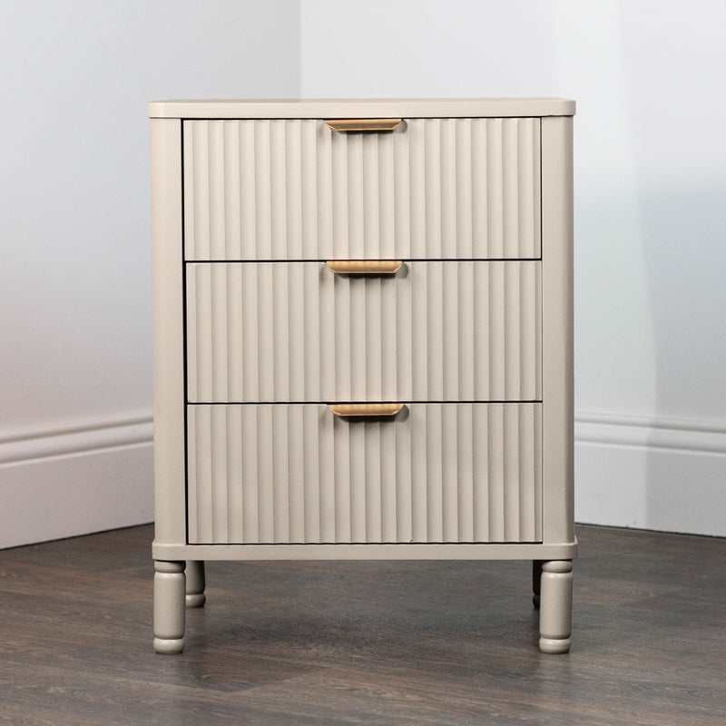 The Parson Locker is a small, light beige three-drawer unit with vertical grooves and gold handles, standing on short legs on a dark wood floor against a white wall.
