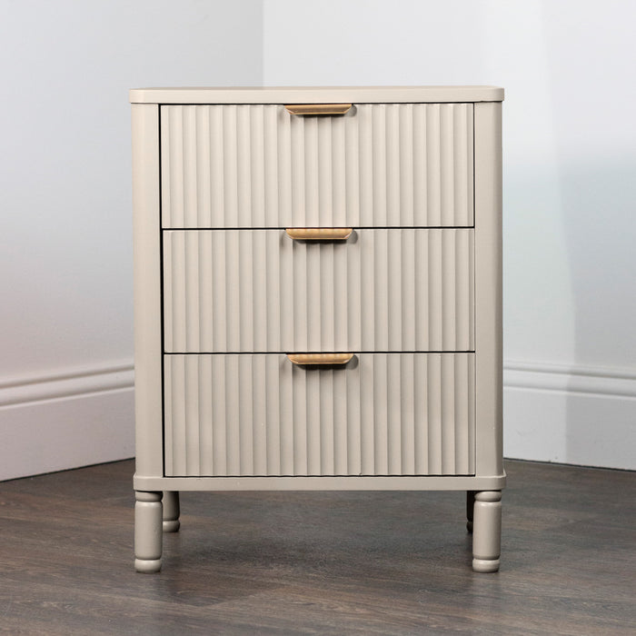 The Parson Locker is a small, light beige three-drawer unit with vertical grooves and gold handles, standing on short legs on a dark wood floor against a white wall.