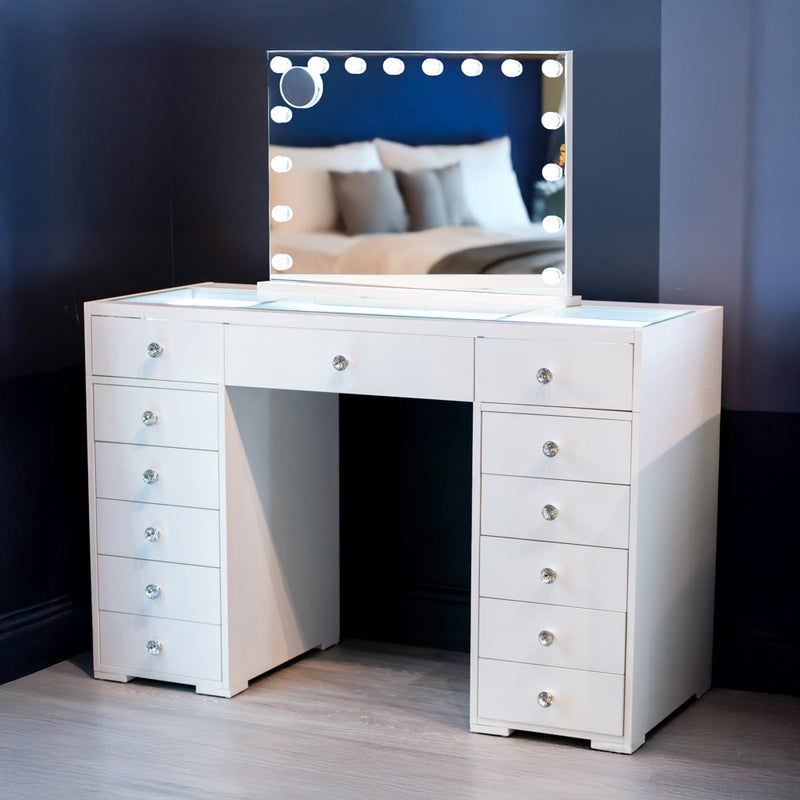 Here’s a rewritten version using your product data: The Erika Hollywood Dressing Table Bluetooth 120cm features a crisp white finish, multiple drawers for storage, and a spacious LED-lit mirror framed by round bulbs. The mirror reflects a softly blurred view of a bed and blue walls in the background.