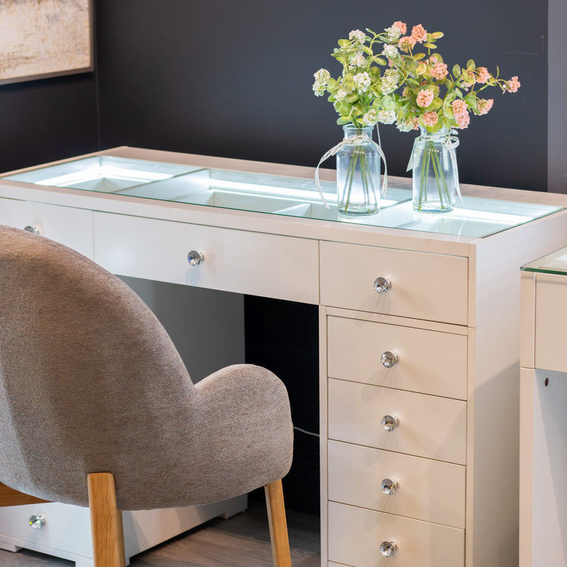 The Erika Hollywood Dressing Table Bluetooth 120cm, with its sleek white finish, glass panels, and chrome handles, is paired with a chic gray upholstered chair to capture the allure of classic Hollywood style. Two clear glass vases filled with pink and white flowers adorn the table, beautifully contrasted against a dark wall backdrop.