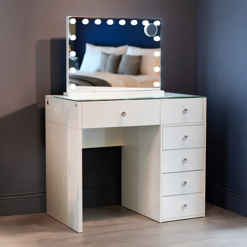 Introducing the Erika Hollywood Dressing Table Bluetooth 92cm: a white vanity with a sleek glass top, five spacious drawers, and a large LED-lit mirror framed by round bulbs. The mirror beautifully reflects a cozy bed with pillows and a blanket in a softly lit room.