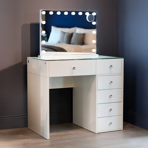 Introducing the Erika Hollywood Dressing Table Bluetooth 92cm: a white vanity with a sleek glass top, five spacious drawers, and a large LED-lit mirror framed by round bulbs. The mirror beautifully reflects a cozy bed with pillows and a blanket in a softly lit room.
