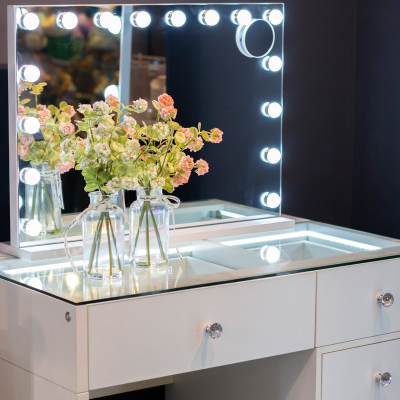 An Erika hollywood dressing table bluetooth 92cm in white, featuring glass drawers, displays two glass vases filled with pink and green flowers in front of a large LED-lit mirror surrounded by round illuminated bulbs.