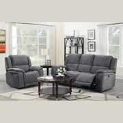 A modern living room features the Boston Recliner Range in dark grey—a corner sofa and matching armchair paired with a round coffee table, decorative shelves, and large windows with white curtains that brighten the space.