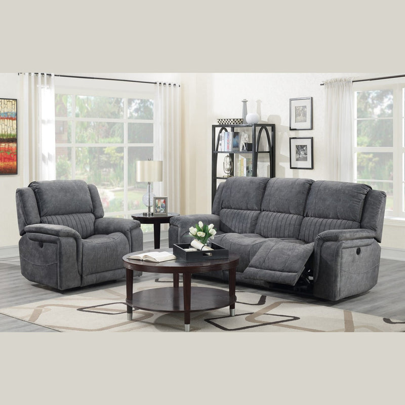 A modern living room features the Boston Recliner Range in dark grey—a corner sofa and matching armchair paired with a round coffee table, decorative shelves, and large windows with white curtains that brighten the space.