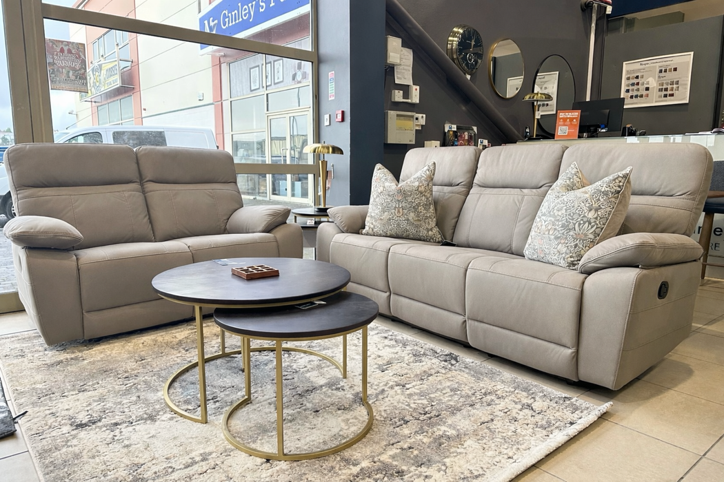 A modern showroom features the Jude Recliner sofa and loveseat in beige washable microfiber with patterned pillows, paired with round nesting coffee tables on a light rug, positioned near large windows and a staircase.