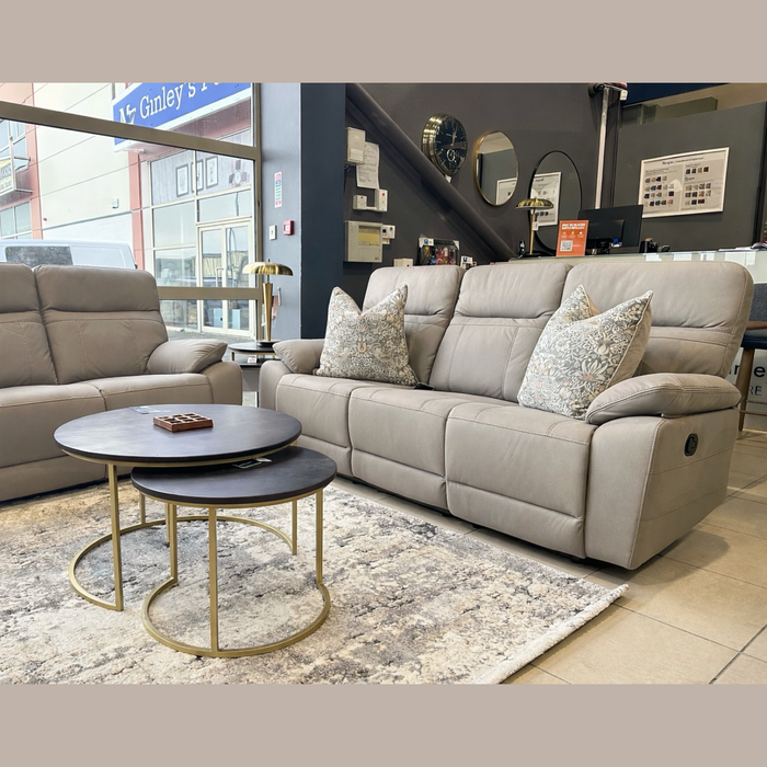 A stylish showroom features a Jude Recliner with patterned pillows, a beige loveseat, a round nested coffee table, and a decorative rug, all arranged near large windows and a staircase.