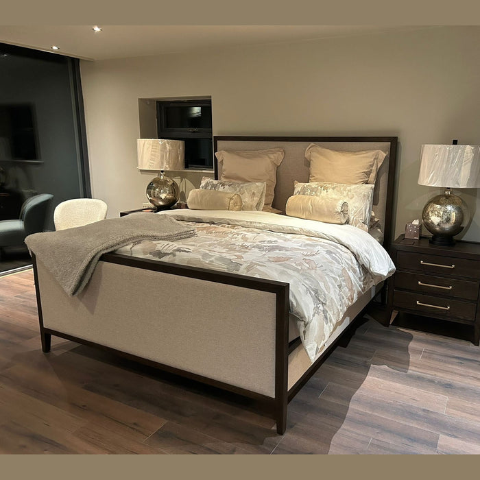 The Hampshire Bedframe, featuring dark wood and luxurious upholstery, is enhanced by a side lamp, bringing elegance to your bedroom.