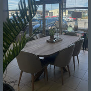 The Alcor Dining Table with six grey chairs is placed near large glass windows overlooking parked cars and buildings. A potted plant, candle holders, and decor are on the table, with a green plant visible in the foreground.