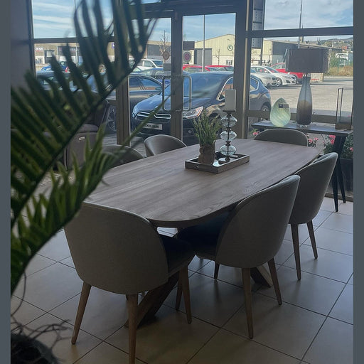 The Alcor Dining Table with six grey chairs is placed near large glass windows overlooking parked cars and buildings. A potted plant, candle holders, and decor are on the table, with a green plant visible in the foreground.