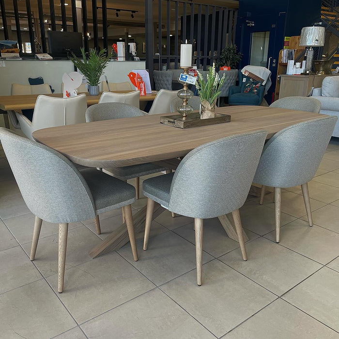 The Alcor Dining Table with a wood finish is surrounded by eight light gray upholstered chairs, adorned with plants, candles, and a tray, and displayed in a furniture showroom with tile flooring.