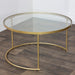 The Andree Coffee Table, with a glass top and elegant brass legs, is displayed on a dark wood floor in a corner with white walls.