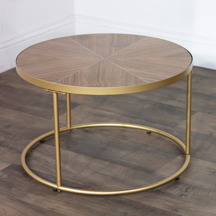 The Andree Coffee Table has a round wood-patterned glass top with elegant brass legs, displayed on a dark wood floor against a white wall.