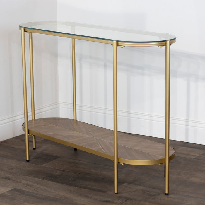 The Andrea Console Table boasts an elegant gold metal frame, oval glass top, and wooden lower shelf with a herringbone pattern—ideal for placement against a white wall on dark wood floors.