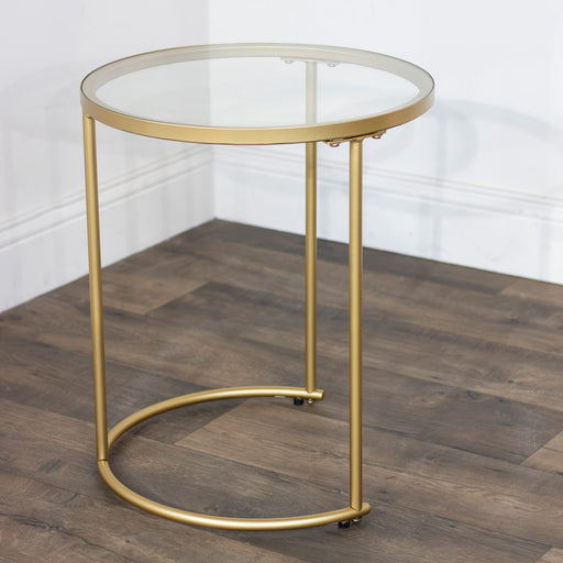 The Andrea Nest of Tables features a small, round design with a geometric gold metal frame and glass top, shown on a wooden floor in the corner of a room with white walls.