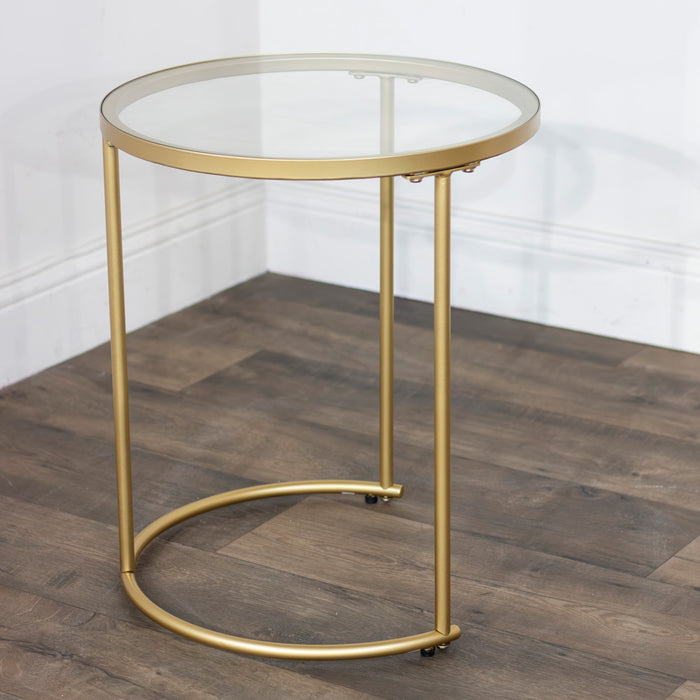 The Andrea Nest of Tables features a small, round design with a geometric gold metal frame and glass top, shown on a wooden floor in the corner of a room with white walls.