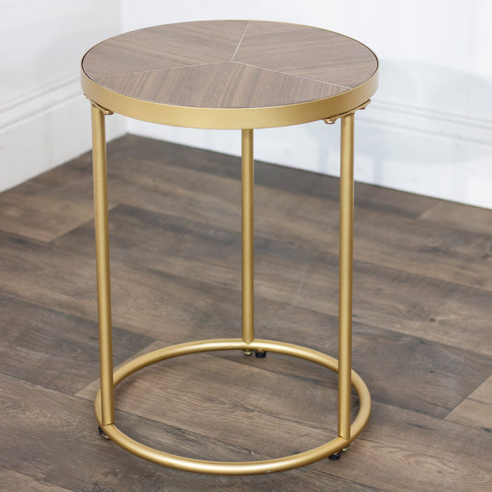 The Andrea Nest of Tables features a small, round tabletop with a wood pattern and gold metal legs in a subtle geometric design, shown on a dark wood floor beside a white wall.