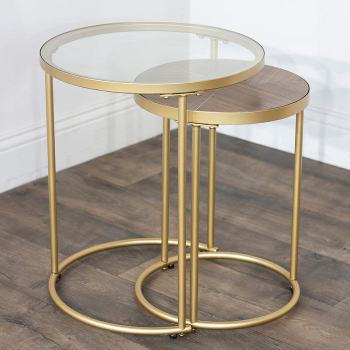 The Andrea Nest of Tables pairs gold metal frames with brass legs; one table features a round glass top, the other a round wooden top. Both are displayed gracefully on a wooden floor against a white wall.