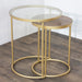 The Andrea Nest of Tables pairs gold metal frames with brass legs; one table features a round glass top, the other a round wooden top. Both are displayed gracefully on a wooden floor against a white wall.