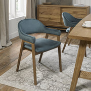 A modern dining room with a wooden table, bright velvet Camden Armchairs, a light-patterned rug, a wooden sideboard, and large curtained windows letting in natural light.