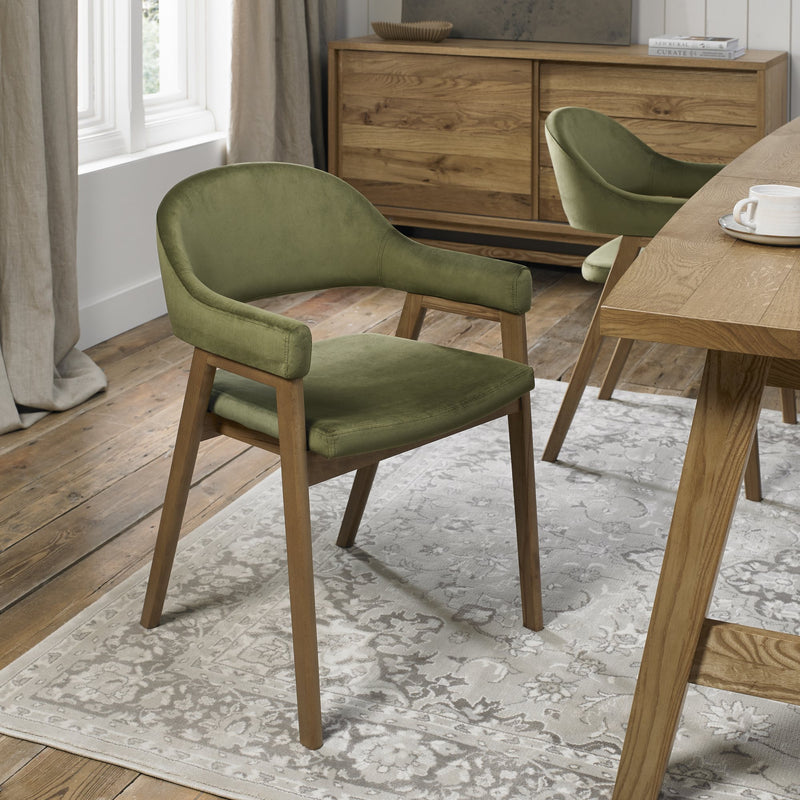A Camden Armchair with a green velvet finish is placed on a light patterned rug beside a wooden dining table in a room featuring wood floors, a sideboard, and large windows with beige curtains.