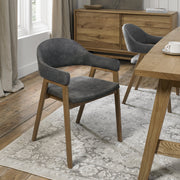 A modern Camden Armchair with wooden legs and a curved, dark gray velvet-upholstered seat sits on a patterned rug in a bright dining room with wooden floors and furniture.