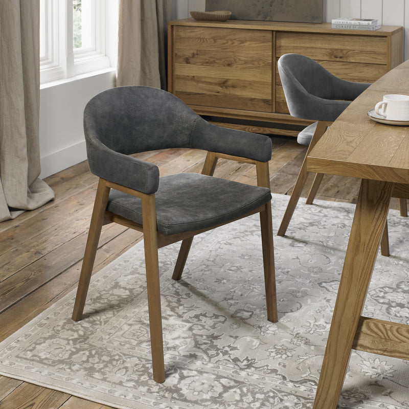 A modern Camden Armchair with wooden legs and a curved, dark gray velvet-upholstered seat sits on a patterned rug in a bright dining room with wooden floors and furniture.