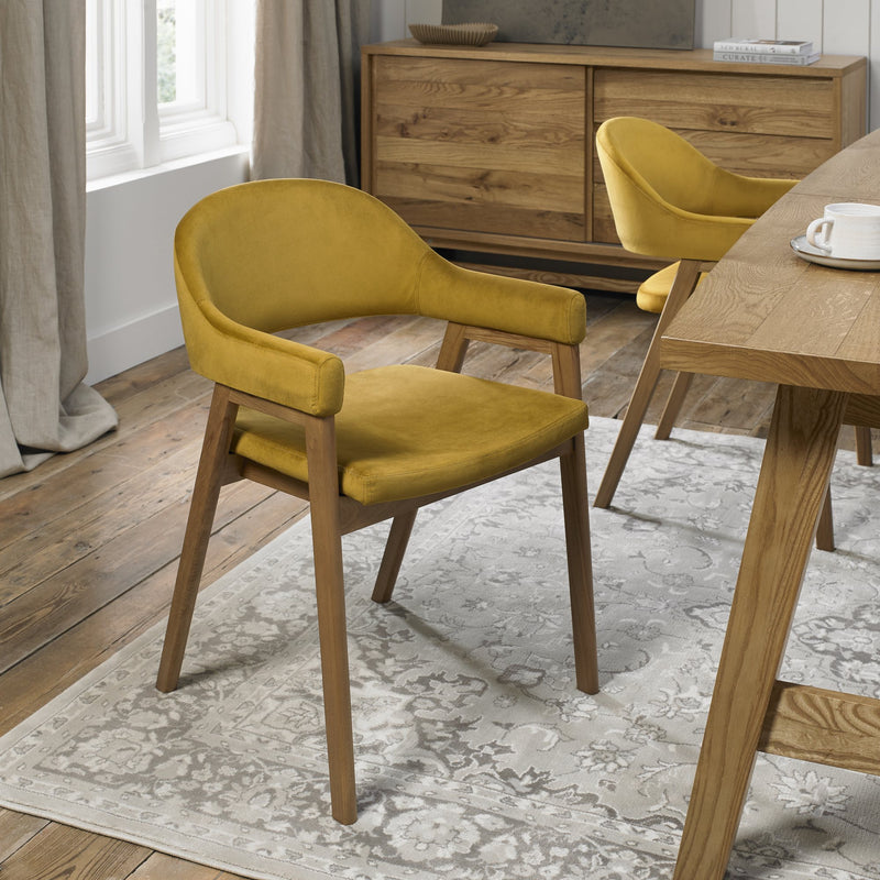 A mustard yellow Camden Armchair with a velvet finish and wooden legs sits on a light patterned rug in a modern dining room with wooden floors, matching furniture, and a table corner holding a mug and plates.