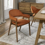 A Camden Armchair with burnt orange velvet upholstery sits on a patterned light gray rug in a dining room featuring wooden floors, a wooden table, and a matching sideboard near a window.