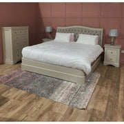 A bedroom with a Mabel Bedframe featuring a fabric headboard, white bedding, two matching nightstands with lamps, a tall dresser, textured area rug, and wood flooring. Muted pink walls have square panel detailing.