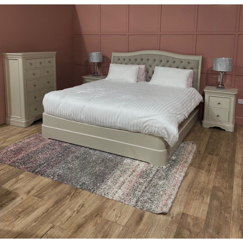 A bedroom with a Mabel Bedframe featuring a fabric headboard, white bedding, two matching nightstands with lamps, a tall dresser, textured area rug, and wood flooring. Muted pink walls have square panel detailing.