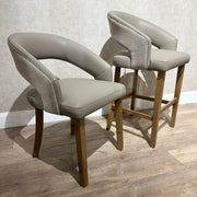 Two Willow Counter Stools with light brown wooden legs, taupe faux leather seats, and textured fabric sides sit on a wood floor in front of a beige textured wall.