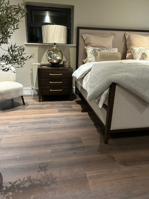 A cozy bedroom showcases the Hampshire Bedframe in dark wood with beige bedding and pillows, a wooden nightstand with a lamp and decorative globe, a cream upholstered chair, a plant, wooden floors, and a window with closed blinds above the nightstand.