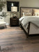 A cozy bedroom showcases the Hampshire Bedframe in dark wood with beige bedding and pillows, a wooden nightstand with a lamp and decorative globe, a cream upholstered chair, a plant, wooden floors, and a window with closed blinds above the nightstand.