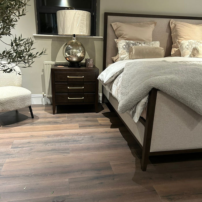 A cozy bedroom showcases the Hampshire Bedframe in dark wood with beige bedding and pillows, a wooden nightstand with a lamp and decorative globe, a cream upholstered chair, a plant, wooden floors, and a window with closed blinds above the nightstand.