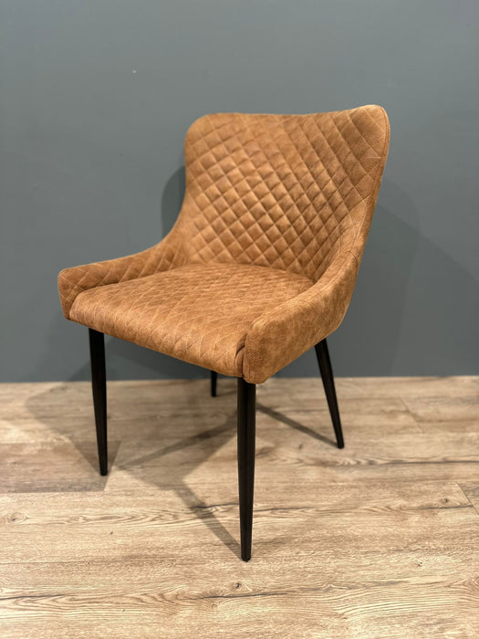 The Diamond Dining Chair, featuring a brown quilted fabric, curved backrest, and black tapered legs, stands on a wooden floor against a plain gray wall.