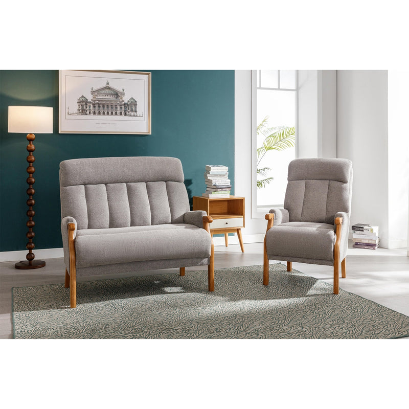 A modern living room with a grey cushioned loveseat, the Blaze Armchair with wooden legs, green accent wall, framed art, floor lamp, small side table holding books, and a patterned rug. Sunlight flows through tall windows.