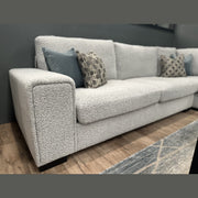 A close-up of the Sorrento Chaise Corner - Special Offer showcases its modern design in light gray neutral textured fabric, complemented by blue and patterned throw pillows. Set against a dark wall and wooden floor, with part of a rug visible in the foreground, this corner sofa offers spacious seating for relaxing in style.