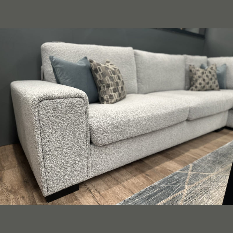 A close-up of the Sorrento Chaise Corner - Special Offer showcases its modern design in light gray neutral textured fabric, complemented by blue and patterned throw pillows. Set against a dark wall and wooden floor, with part of a rug visible in the foreground, this corner sofa offers spacious seating for relaxing in style.