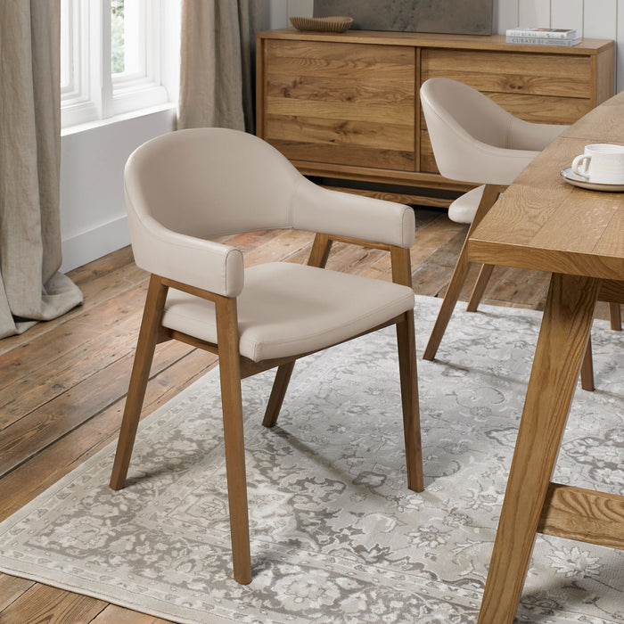 A Camden Armchair with light beige upholstery and wooden legs is placed on a patterned rug in a dining room featuring wood flooring, a wooden table, and a sideboard by the window.