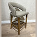 The Willow Counter Stool features a modern counter-height design with a light wood frame, tan cushioned seat, and curved backrest upholstered in smooth and textured beige fabric, shown against a light wall and wood floor.