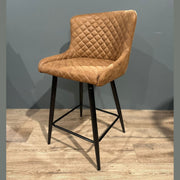 The Diamond Counter Stool features a modern brown PU leather, diamond-quilted seat and backrest, black metal legs with footrest—ideal for kitchen islands, shown on a light wood floor against a gray wall.