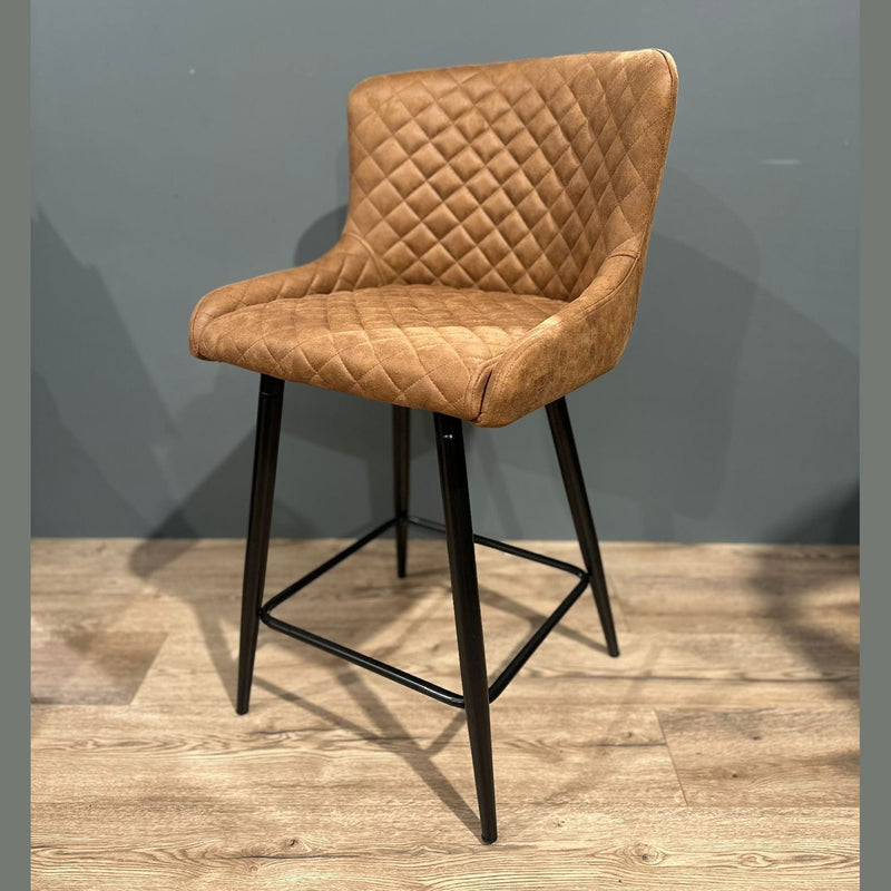 The Diamond Counter Stool features a modern brown PU leather, diamond-quilted seat and backrest, black metal legs with footrest—ideal for kitchen islands, shown on a light wood floor against a gray wall.