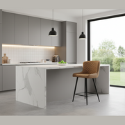 A modern kitchen with gray cabinets, a marble island, and a Diamond Counter Stool with quilted diamond stitching in brown PU leather. Two black pendant lights hang above the island, while large glass doors showcase a lush outdoor view.