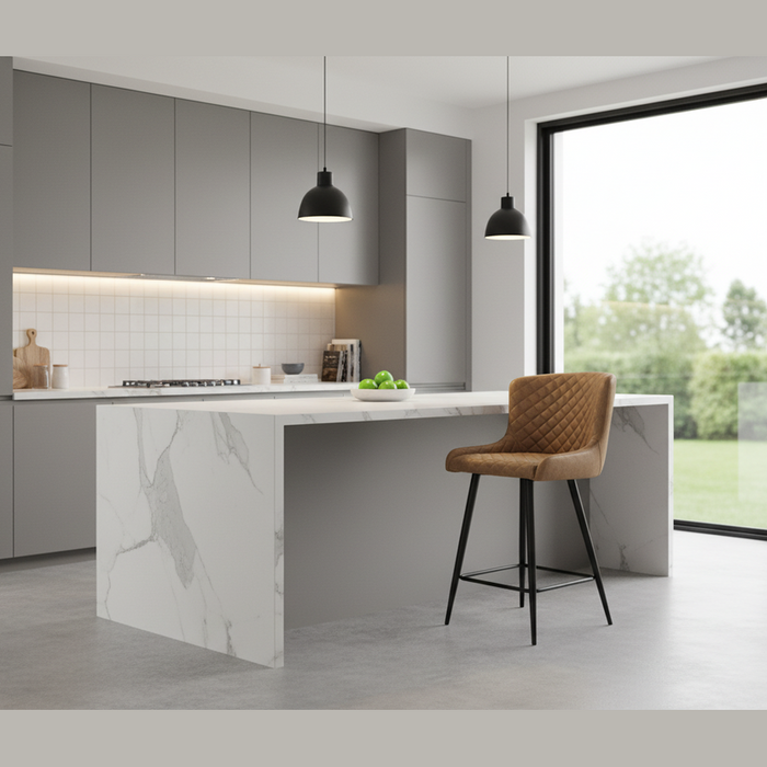 A modern kitchen with gray cabinets, a marble island, and a Diamond Counter Stool with quilted diamond stitching in brown PU leather. Two black pendant lights hang above the island, while large glass doors showcase a lush outdoor view.