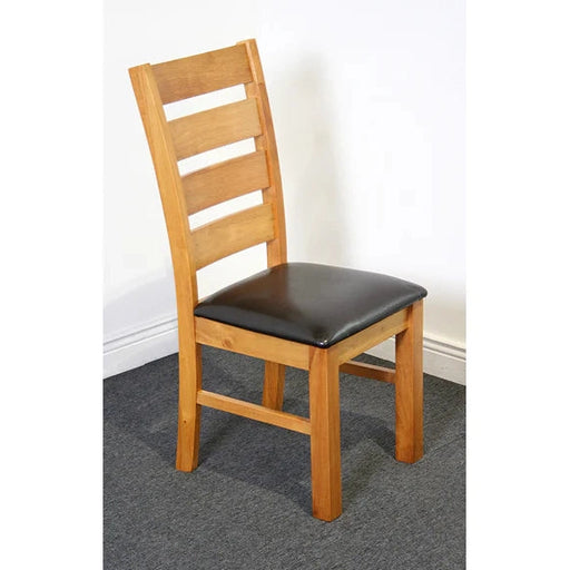 A wooden dining chair with a ladder-back design and black padded seat from the Columbia Dining Set - Oak, placed on a gray carpet against a white wall.