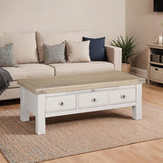 The Doune Coffee Table in light oak veneer with three storage drawers sits on a woven rug in front of a beige sofa with patterned cushions. A plant is beside the sofa and a cabinet with baskets can be seen in the background.