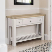 The Doune Console Table is a white table with a light wood top, three drawers, and an open slatted shelf. It stands on tiled flooring by a cream wall and doorway.