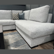 The Sorrento Chaise Corner - Special Offer, upholstered in light gray neutral textured fabric and featuring two decorative pillows, provides ample seating atop a patterned rug beside a dark wooden coffee table in a modern living room with gray walls and wood flooring.