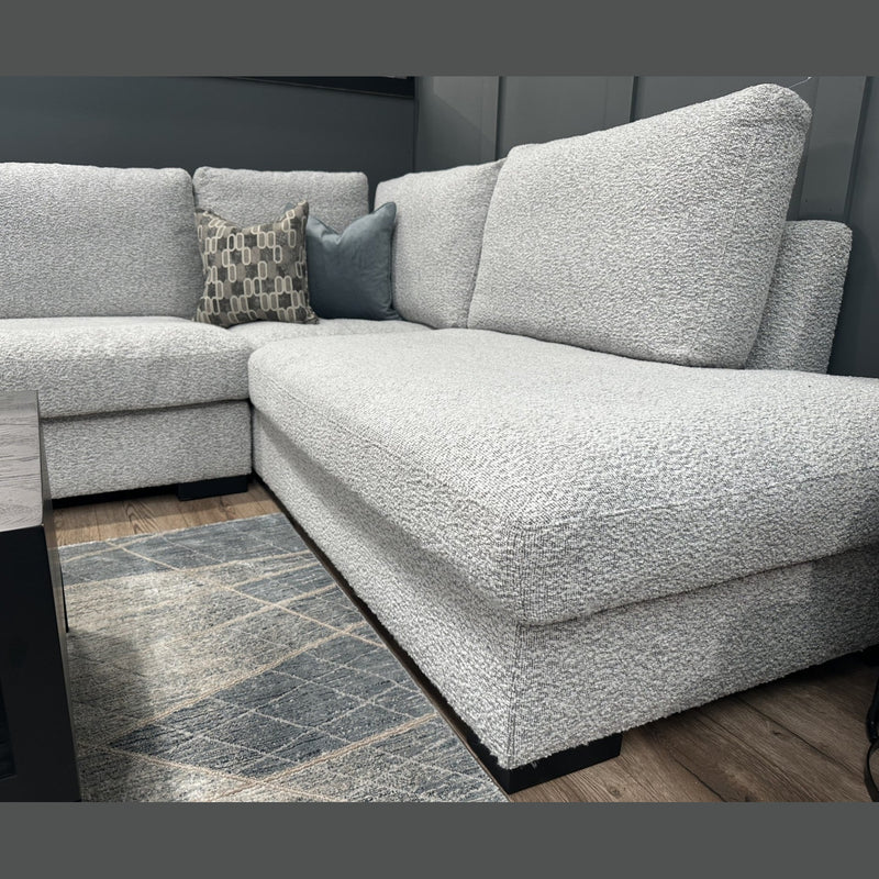 The Sorrento Chaise Corner - Special Offer, upholstered in light gray neutral textured fabric and featuring two decorative pillows, provides ample seating atop a patterned rug beside a dark wooden coffee table in a modern living room with gray walls and wood flooring.